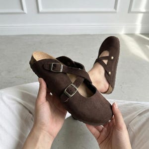 Könnte beinhalten: Ein Paar dunkelbraune Wildleder-Clogs mit abgerundeter Zehenpartie, einem Fußbett in Korkoptik und zwei verstellbaren Riemen mit Metallschnallen auf der Oberseite. Das Design umfasst gekreuzte Riemen und eine praktische Schlupfform.