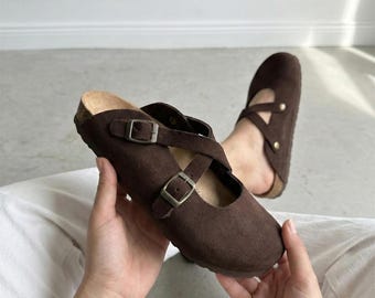 May include: A pair of dark brown suede clogs featuring a rounded toe, a cork-style footbed, and two adjustable straps across the top with metallic buckles. The design includes a crisscross strap detail and a slip-on style construction.