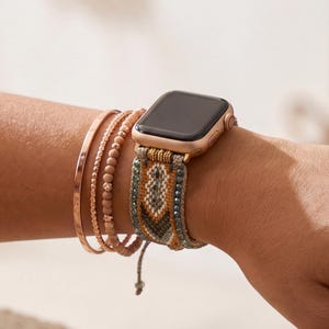 Könnte beinhalten: Eine goldene Apple Watch mit einem gemusterten Armband in Grün-, Braun- und Blautönen wird am Handgelenk getragen, zusammen mit mehreren roségoldenen Armbändern. Die Armbänder umfassen einen gehämmerten Armreif, Perlenstränge und eine zarte Kette.