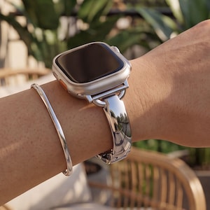May include: A silver-toned metal watch strap with a polished finish attached to a rectangular smartwatch with a light-coloured casing and an orange side button, accompanied by a thin, smooth silver-toned metal bangle bracelet on the same wrist.