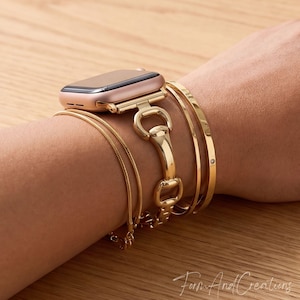May include: A gold Apple Watch with a matching strap and multiple gold bracelets. The watch strap has an equestrian-inspired design. The bracelets include a fine chain, a solid band, and a band with a small gem. The items are displayed on a light-coloured wooden surface.