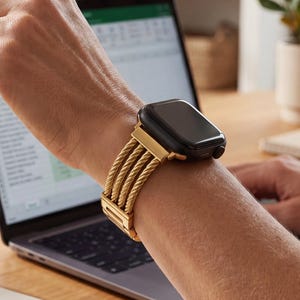 May include: A gold-toned metal watch strap featuring four parallel twisted rope-style strands attached to a black smartwatch face. The strap includes a secure metal clasp mechanism and connects directly to the watch housing with a polished gold finish.