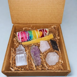 Puede incluir: Una caja de cartón marrón llena de varios cristales, un manojo de salvia envuelto en hilo arcoíris y palo santo. Los cristales incluyen amatista, cuarzo rosa y selenita. La caja está forrada con papel de seda marrón.