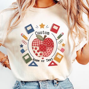 以下が含まれることがあります： アイボリーのTシャツ。中央にはパッチワーク風のリンゴのグラフィックが描かれ、その周りにノート、鉛筆、ペーパークリップ、消しゴム、定規などの学用品が配置されています。リンゴの周りには「Custom Name or Text」という文字がプリントされています。