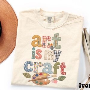 May include: An ivory short-sleeve t-shirt featuring the text "art is my Craft" in a patchwork-style font with various floral, gingham, and solid patterns. Below the text is a wooden paint palette with colorful paint dabs and a paintbrush.