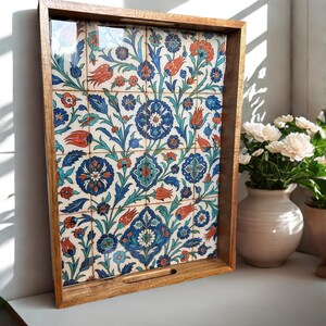 Op de afbeelding: Houten dienblad met een decoratief bloemen tegelinzetstuk. De tegel heeft een patroon van blauwe, oranje en teal bloemen en bladeren op een witte achtergrond. De lade heeft een lichtbruin houten frame.