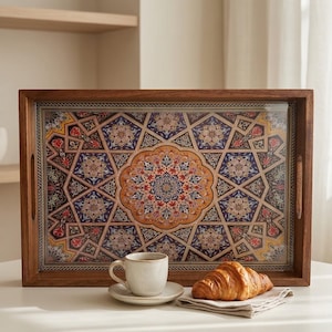 Könnte beinhalten: Ein rechteckiges Serviertablett aus Holz mit einem farbenfrohen geometrischen Muster. Das Tablett hat einen dunkelbraunen Rahmen und integrierte Griffe. Eine Tasse Kaffee und ein Croissant liegen auf dem Tablett, was auf ein Frühstück hindeutet.