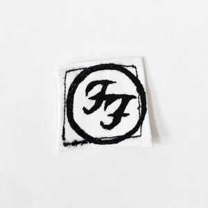 May include: A square white fabric patch featuring a black embroidered design. The design consists of a hand-stitched black circle enclosing the stylised black letters FF, which are also hand-stitched onto the white fabric surface.