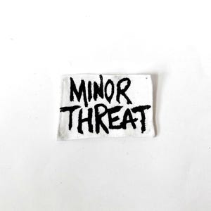 May include: A rectangular white fabric patch featuring the text MINOR THREAT printed in a bold, black, hand-drawn style font. The lettering has a distressed, ink-like appearance with uneven edges, centred on the white background of the patch.