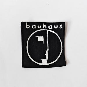 May include: A square black fabric patch featuring the white embroidered text "bauhaus" at the top. Below the text is a white circular outline containing a stylised, abstract profile of a face, rendered in bold white lines against the black background.