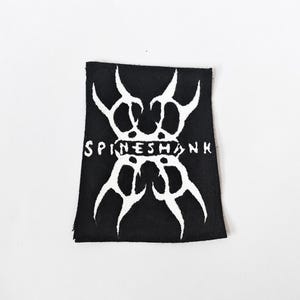 May include: A rectangular black fabric patch featuring a white embroidered graphic design. The design consists of symmetrical, sharp, thorn-like shapes arranged vertically with the word SPINESHANK printed in white capital letters across the centre.