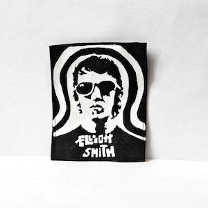 May include: A rectangular black and white fabric patch featuring a high-contrast graphic illustration of a person wearing sunglasses with a curved line design behind the head and the text Elliot Smith printed in white lettering at the bottom.