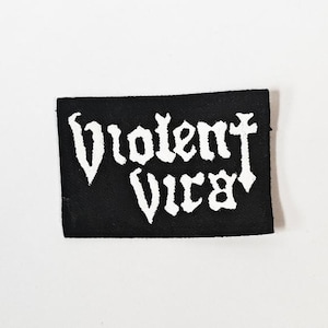 May include: A rectangular black fabric patch featuring the white embroidered text Violent Vira in a stylised, jagged font. The patch has a clean, finished edge and is presented against a plain, neutral background, highlighting the contrast.