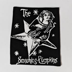May include: A rectangular black fabric patch featuring a white screen-printed graphic of a person sitting on a large star surrounded by small star shapes. The text "The" appears at the top left, and "Smashing Pumpkins" is printed at the bottom.