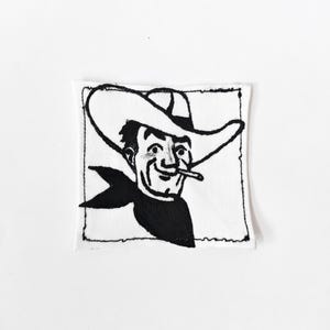 May include: A square white fabric patch featuring a black ink illustration of a smiling person wearing a wide-brimmed cowboy hat and a neckerchief. The person has a thin object held between their lips. The design is enclosed in a black border.