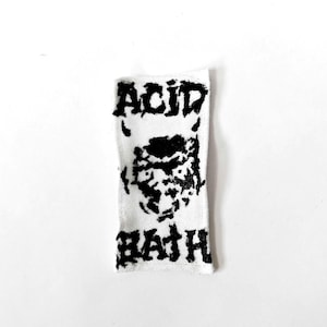 May include: A rectangular white fabric patch featuring a black screen-printed graphic. The design includes the text ACID at the top and BATH at the bottom, with a stylised, distressed illustration of a face with horns positioned in the centre.