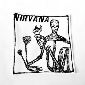 May include: A square fabric patch featuring a black ink screen print on a white background. The design includes the text NIRVANA at the top, with a central illustration of a tall, thin figure, a small horned creature, and two flower-like shapes.