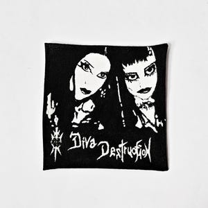 May include: A square black and white fabric patch featuring a high-contrast graphic illustration of two faces with dark makeup. The text Diva Destruction is printed in a stylised font at the bottom, accompanied by a small heart and dagger icon.