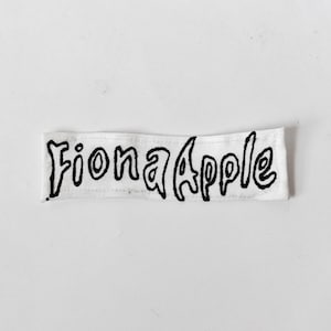 May include: A rectangular white fabric patch featuring the text Fiona Apple embroidered or printed in a bold, black, hand-drawn style font. The patch has visible stitching along the top and bottom edges, creating a clean, finished look for application.