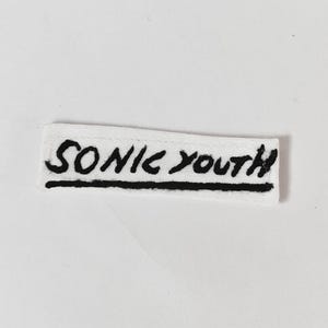 May include: A rectangular white fabric patch featuring the text SONIC YOUTH embroidered in black thread. The lettering has a hand-drawn, slightly irregular style, and a solid black horizontal line is stitched directly underneath the band name.