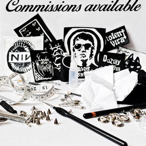May include: A collection of black and white fabric patches featuring various graphic designs and text, including 'NIV', 'London After Midnight', and 'Dazey'. The text 'Commissions available' appears at the top. Tools include a measuring tape, safety pins, metal studs, a black crochet hook, and a small plastic bottle.