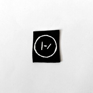 May include: A square black fabric patch featuring a white embroidered circular emblem. Inside the circle, there is a vertical line on the left and a diagonal slash on the right, forming a stylised logo design with clean, contrasting white stitching.