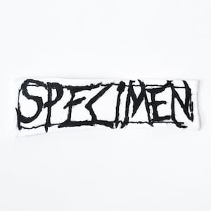 May include: A rectangular white fabric patch featuring the word SPECIMEN embroidered in a bold, jagged, black font. The lettering has a distressed, hand-drawn appearance with uneven lines and sharp edges, centred on the white rectangular textile base.