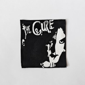 May include: A square fabric patch featuring a high-contrast black and white graphic. The design displays the text THE CURE in a stylised font at the top, accompanied by a partial, shadowed portrait of a face on the right side of the patch.