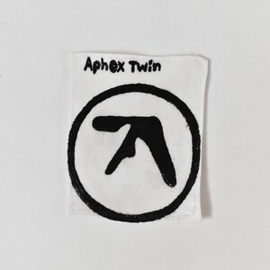 May include: A rectangular white fabric patch featuring a hand-drawn black Aphex Twin logo centred within a black circle. The text Aphex Twin is written in black ink at the top of the patch. The edges of the white fabric are stitched with white thread.
