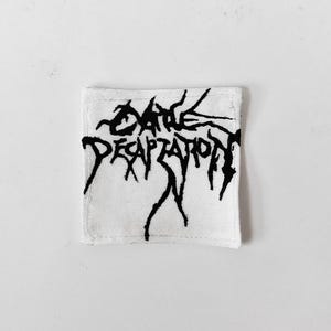 May include: A square white fabric patch featuring the black embroidered text Cattle Decapitation in a jagged, stylised font. The patch has visible stitching along its four edges, creating a clean border around the central graphic design.