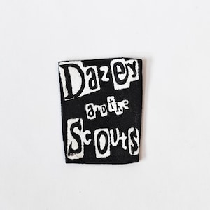 May include: A rectangular black fabric patch featuring the white hand-drawn text Dazey and the Scouts. The lettering is stylised with a bold, uneven font, and the patch has a visible woven texture throughout the black background material.