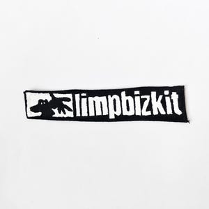 May include: A rectangular black fabric patch featuring a white embroidered graphic of a stylised hand on the left side and the text limpbizkit in white lettering on the right side. The patch has a clean, straight-edged border design.
