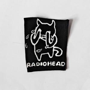 May include: A rectangular black fabric patch featuring a white embroidered graphic of a crying creature with pointed ears and a tail. The word RADIOHEAD is embroidered in white capital letters at the bottom of the patch design.