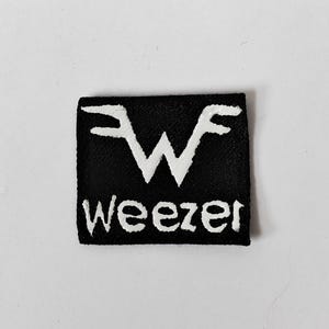 May include: A square black fabric patch featuring the white embroidered Weezer logo. The design includes a stylised letter W symbol at the top and the word Weezer written in white lowercase letters underneath on a solid black background.