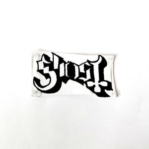 May include: A rectangular fabric patch featuring a white background with the word Ghost printed in a bold, black, stylised gothic font. The lettering has a distinct drop shadow effect, and the edges of the patch are finished with white stitching.