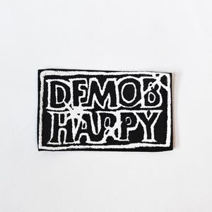 May include: A rectangular embroidered patch featuring a black background with a white border and the text DEMOB HAPPY in a stylised, bold font. The lettering is outlined in white, creating a high-contrast graphic design on the fabric surface.