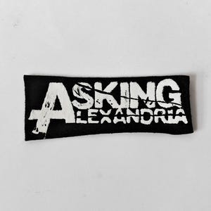May include: A rectangular black fabric patch featuring the white text Asking Alexandria in a distressed, weathered font style. The lettering is bold and occupies the majority of the patch surface, creating a high-contrast visual design.