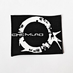May include: A rectangular black fabric patch featuring a white graphic design. The design consists of a circular, distressed ring shape with a jagged, star-like extension on the right side. The text CHEMLAB is printed in white across the centre.