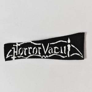 May include: A rectangular black fabric patch featuring white embroidered text that reads Horror Vacui. The lettering is stylised with sharp, angular edges and a pointed design element on the right side, creating a high-contrast graphic appearance.