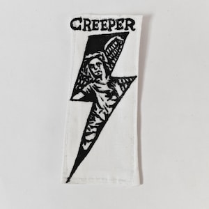May include: A rectangular white fabric patch featuring a black screen-printed design of a lightning bolt containing a figure with wings. The word CREEPER is printed in black capital letters at the top of the patch above the lightning bolt graphic.