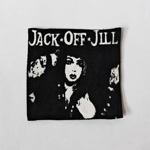 May include: A square black and white fabric patch featuring the text JACK OFF JILL at the top in a stylised font. Below the text is a high-contrast graphic illustration of a person with dark hair and distinct facial features centred on the patch.
