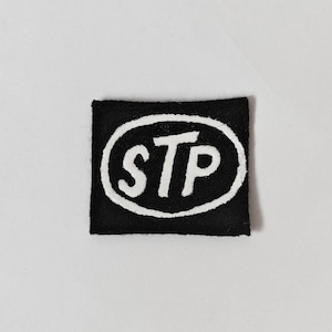 May include: A square-shaped black fabric patch featuring a white embroidered oval border and the letters STP in white, centred within the oval. The design is stitched onto a black background, creating a high-contrast graphic emblem for clothing.