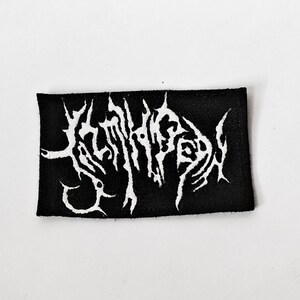 May include: A rectangular black fabric patch featuring white embroidered text in a stylised, jagged, and illegible font. The design consists of sharp, pointed lines and intricate, thorny shapes that span the entire width of the black background.