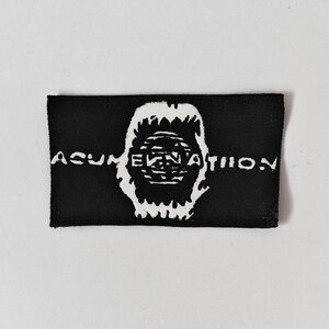 May include: A rectangular black fabric patch featuring a white graphic design in the centre. The design depicts a stylised, jagged-edged circular shape with horizontal lines inside, flanked by the white text ACUMEN NATION on both sides.