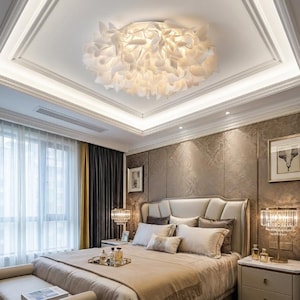 May include: A bedroom featuring a large, white, floral-shaped ceiling light fixture. The room has a bed with a beige comforter, decorative pillows, and two bedside lamps. The walls are a textured, neutral color, and the room has curtains.