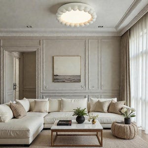 May include: A contemporary living room featuring a white corner sofa, a marble-topped coffee table, and a decorative ottoman. A large, round, white ceiling light with a scalloped edge illuminates the space. The walls are painted a neutral colour, and a painting hangs above the sofa.