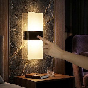 May include: A modern wall sconce with a rectangular frosted glass design, featuring a black band in the center. The sconce is mounted on a dark marble-patterned wall. A hand is touching the light. A small table with a glass and a book is in the foreground.
