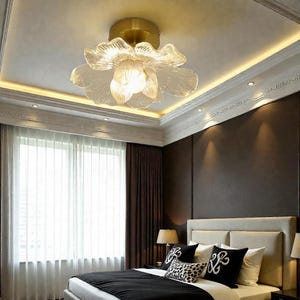 Puede incluir: Una lámpara de techo con una pantalla de cristal en forma de flor. La lámpara tiene una base dorada y una fuente de luz brillante. La habitación incluye una cama con almohadas y una ventana con cortinas.