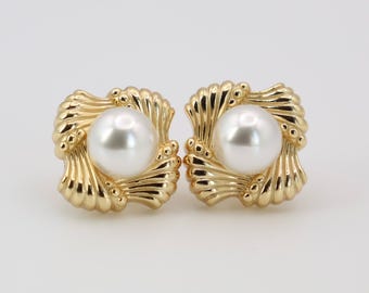 May include: A pair of gold-colored square stud earrings. Each earring features a large, round, white pearl set within a decorative gold frame with a shell-like design. The earrings are displayed against a plain white background.