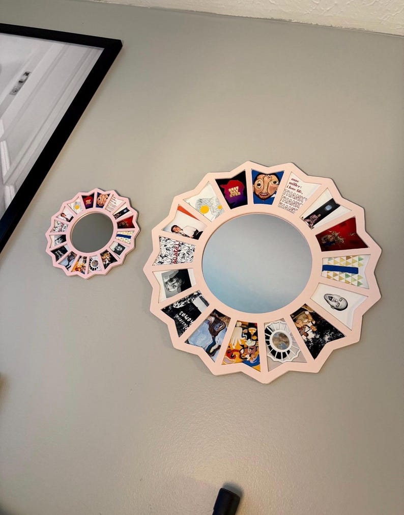 May include: Two circular wall mirrors with light pink frames featuring a scalloped, star-like edge. Each mirror is surrounded by a series of small, square, multi-coloured graphic art prints arranged in a radial pattern around the central glass.